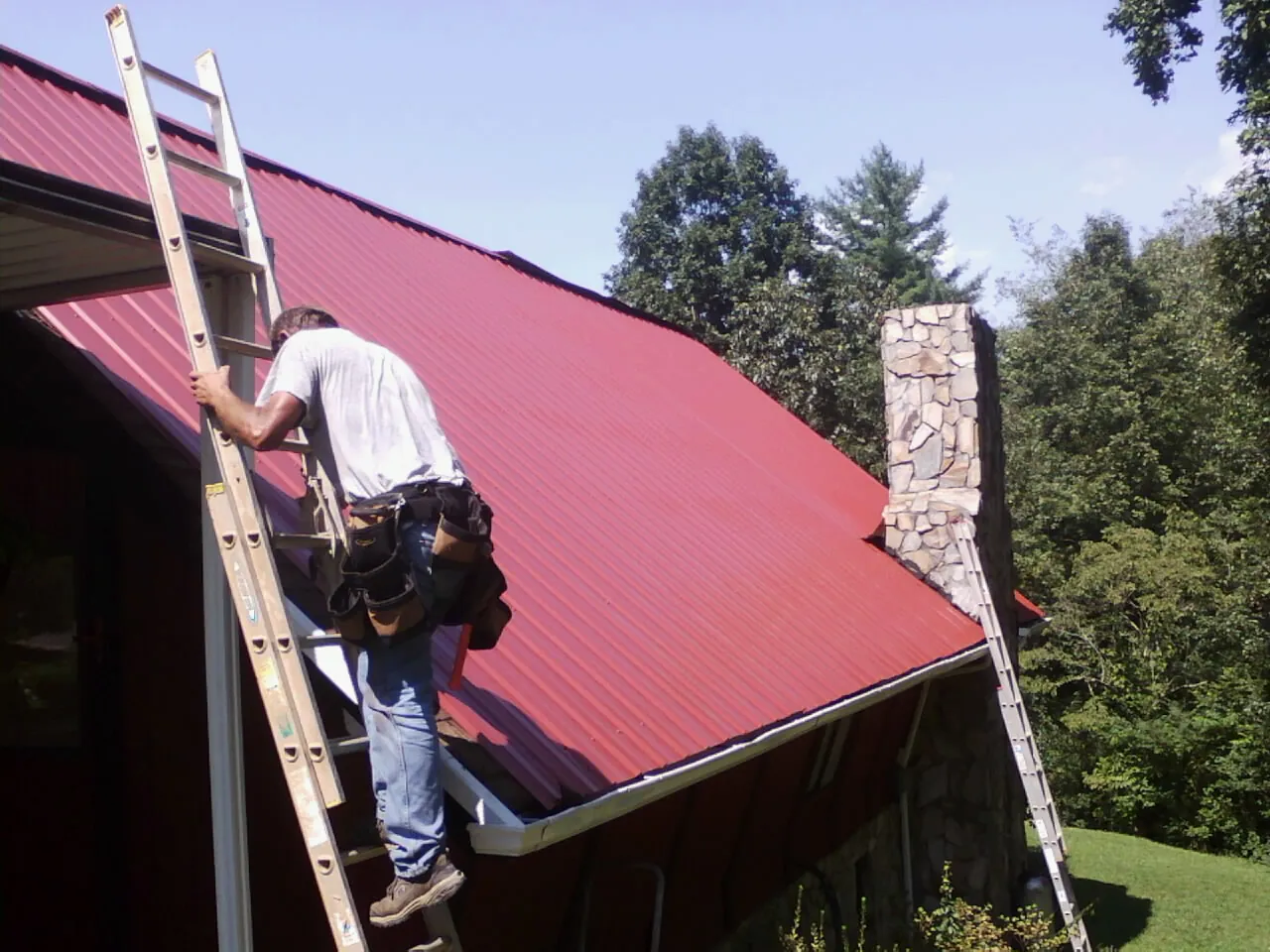Roofer on a ladder beside a red metal roof and stone chimney — Soffit & Fascia Repair by Roofing Brothers in Sharps
